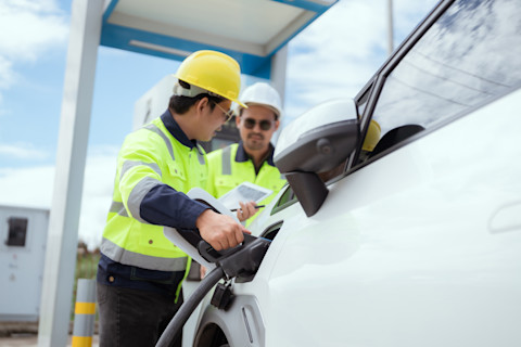 Two workers in safety gear are engaged in setting up charging equipment for an electric vehicle at a modern charging station under clear skies and minimal cloud cover.