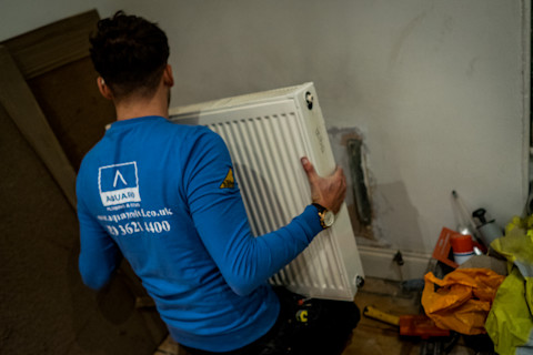 A tradesperson from the company 'AQUAID' is installing a white radiator on a wall in an indoor setting. The individual, seen from the back, is wearing a blue company shirt with a logo and contact information on the back, along with protective gloves. The radiator is being held up against a partially prepped wall, with some construction debris and tools scattered on the floor, indicating an ongoing installation or renovation process.