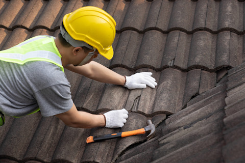This image shows a roofing specialist wearing a high vis vest and hard hat. He is working on fixing roof tiles. There is a hammer to the right of him.
