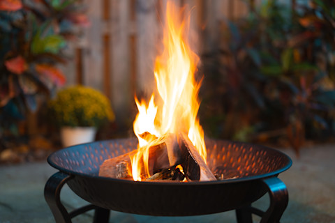 A small black metal firepit on a patio in a back garden with a fire lit within, with a brown fence and several green plants in the background.
