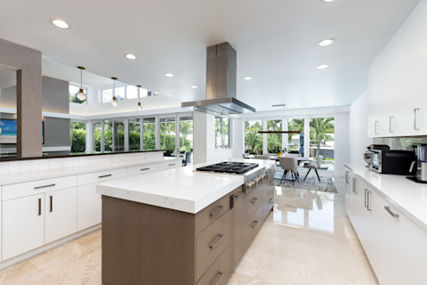 This image shows a bright and modern kitchen with a large kitchen island in the centre of the room. The cabinets and worktops around the edge of the room are white and the island's cabinets are brown. The floor has glossy beige tiles and there is a window at the bottom of the room overlooking green vegetation.