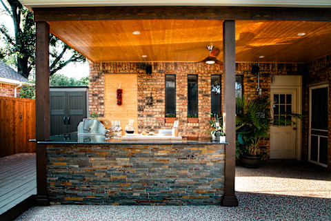A stylish outdoor kitchen featuring a sleek countertop with a built-in grill and various kitchen utensils. The kitchen is set against a backdrop of a brick wall with decorative windows and a wooden ceiling, creating a cozy and inviting space. The area is well-lit, and the surroundings include a wooden deck, potted plants, and a storage shed, emphasizing a well-designed and functional outdoor cooking and dining area. The image captures the essence of a high-quality outdoor kitchen setup.