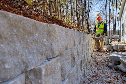 A large, stone garden wall under construction. Already half built, the wall stands 3 stones deep, and a construction worker in a hi-vis and a hard hat can be seen at the far end of the wall adding a new brick to the wall.