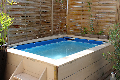 A wooden framed pool with wood steps set up at the bottom of a garden in front of a wooden fence and surrounded by tropical plants.