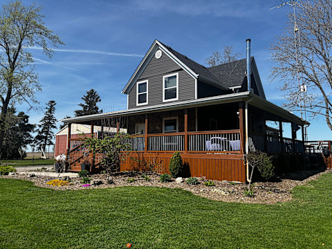 Charming two-story house with grey siding and a spacious wooden, wrap around extension porch. Surrounded by green lawn, garden beds, and tall trees under a clear blue sky.