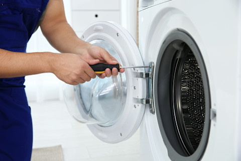 A close-up of a technician in blue work clothes repairing a washing machine. The person is holding a screwdriver and working on the hinges of the front-loading washing machine's door. The door is open, revealing the washing machine's drum. The screwdriver is being used to adjust or remove the screws on the door. The environment is bright, and blurred white furniture can be seen in the background.