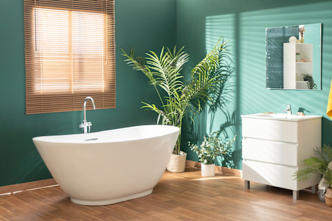 This image shows a modern, brightly lit bathroom featuring green walls, a white freestanding bath, leafy plants and light wood accents.