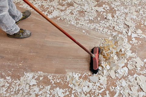 This image shows a cleaner or tradesperson using a brush to sweep a laminate floor that is covered with building residue after a house renovation. The cleaner or tradesperson is getting the room back to a liveable state.