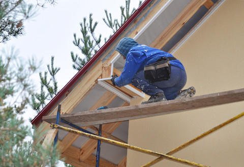 Close-up of a plaster cornice being installed on a ceiling. The image shows a technician applying and smoothing plaster around the cornice, with scaffolding or a ladder visible in the background. The surrounding ceiling and walls are partially finished.