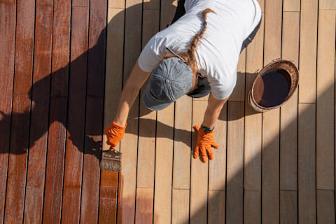 A handyman using a dark brown stain on light brown decking boards.