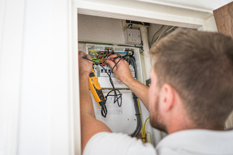 This image shows a pair of hands installing an electric fuse box on the wall of a house or commercial building. The wall is unfinished; it still needs to be painted or wallpapered.