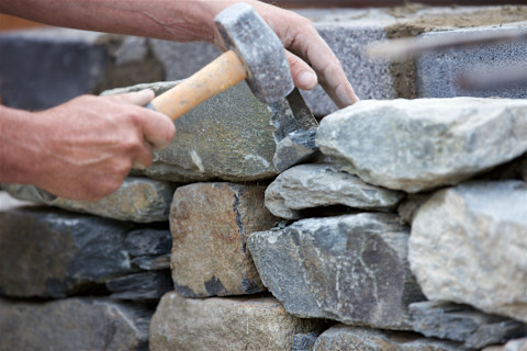 Close‑up of a stonemason’s hands using a hammer and chisel to shape and fit stones in a dry stone wall construction.
