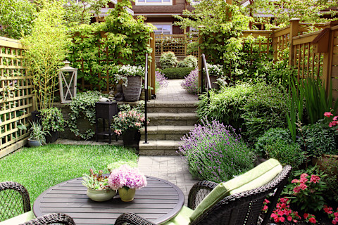 This image shows a small tiered garden with a seating area and a small patch of lush green grass. On both sides of the garden there are a lot of plants and flowers.
