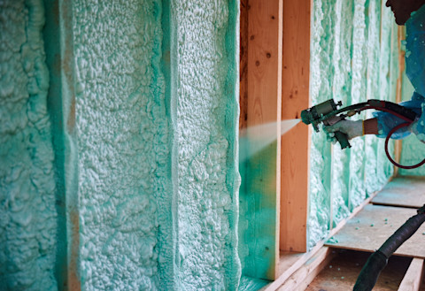 A worker sprays green foam insulation onto wooden wall studs inside a building under construction, creating a thick and uneven texture.