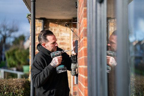 An electrician standing in a porch of a building with a drill in hand fixing an outdoor socket.