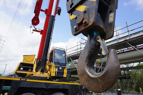 A large mobile crane for construction with a yellow cabin and red boom, featuring a rugged metal hook in the foreground. Scaffolding and a blue sky with clouds are in the background.