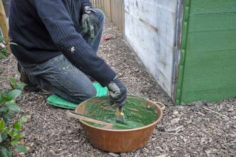 This image shows a hand painting a wooden garden shed with green paint using a brush. The person is kneeling on a pad and is wearing outdoor gloves to project their hands.