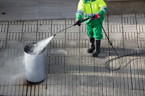 A professional bin washing service in action, showing a worker cleaning a large outdoor waste bin using a high-pressure washing machine. The setup includes a specialized vehicle equipped with cleaning tools, highlighting the bin washing process and the professional equipment used to maintain hygiene.