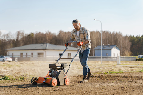 Close-up of a lawn aeration service in progress. A worker is using a core aerator machine to punch holes into a grassy lawn, with soil plugs visible on the surface. The background shows a well-maintained yard with green grass and trees