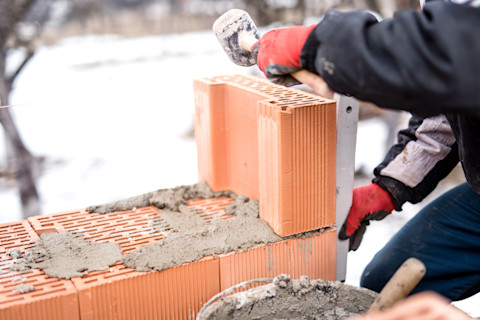 This image shows a mason or bricklayer working on a snowy construction site, carefully placing bricks and applying mortar to build a sturdy wall.