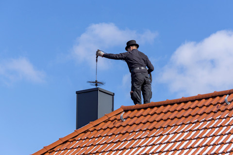 This image shows a modern chimney sweep standing on a terracotta roof top of a residential house. He is preparing to send a brush on a cable down the chimney.