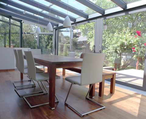 This image shows a bright conservatory being used as a dining room. There is a dining table and chairs and wooden flooring. Through the panes of glass you can see a green garden with leafy green trees.