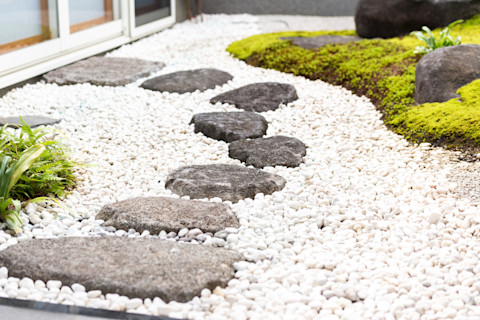 A garden path, made up of smooth black stones, that leads across a gravel lawn. The gravel is bordered by moss and rocks, and leads to a patio door.