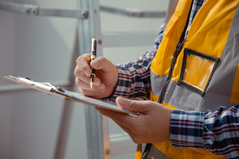 This image shows a building contractor inspecting a construction site and writing down details on a clipboard. The contractor is wearing a reflective vest.