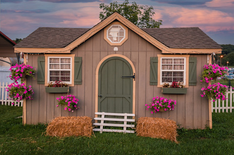 This image shows a garden shed with a shingle roof, windows, hanging baskets with pink flowers and hay bales by the door.