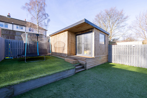A large garden office building on a raised deck in a backyard, beside a trampoline and a fence.