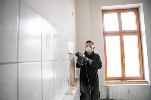 A construction worker wearing protective goggles and a face mask uses a power tool to remove tiles from a wall in a brightly lit room with large windows.