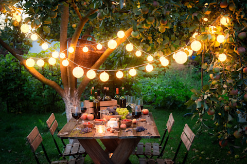 This image shows an outdoor table in a garden with food and drinks at night time beneath glowing string lights.