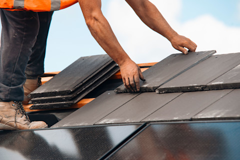 A construction worker installs dark roof tiles on a rooftop under a clear blue sky. They wear work boots and an orange safety vest.