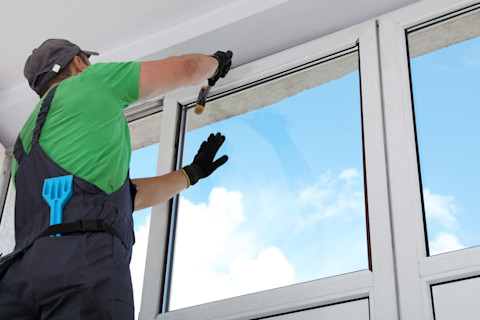 A worker in a green shirt and black overalls installs a large window, using a hammer. A bright blue sky with clouds is visible through the glass.