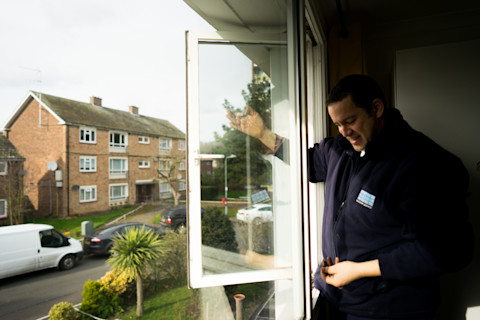 An individual in a navy blue work fleece with a visible company logo on the chest is engaged in window maintenance indoors. They appear to be adjusting or examining the hinge of an open white-framed window that overlooks a residential area with brick apartment buildings and parked cars. The setting suggests a routine window servicing task in an urban neighborhood during daylight, with soft natural light entering through the window.