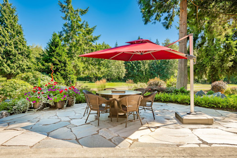 The image shows a paved patio with an outdoor dining table shaded by a red sun shade. There are plants, shrubs, trees and green grass in the background and a cloudless blue sky.