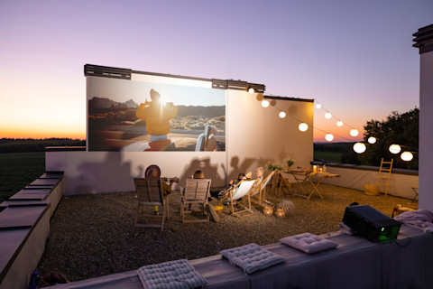 People sitting on chairs in a garden watching a movie. There are lights and some cushions as well.