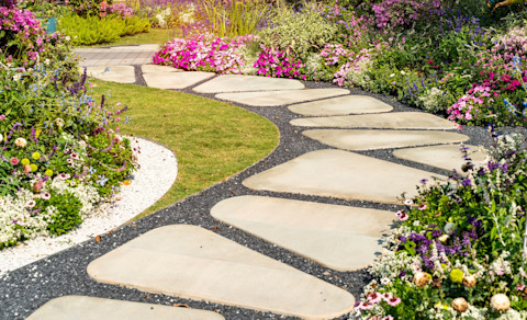 This image shows a stone and gravel path winding through a lush garden with green shrubs and blooming flowers on either side, creating a peaceful outdoor walkway.
