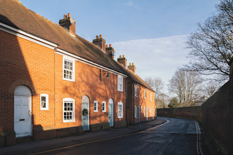 A row of traditional red-brick terraced houses on a quiet, curved street. The houses feature white-framed windows and doors, some of which have decorative wreaths. The brickwork and chimneys reflect the charm of classic British architecture, with arched doorways adding to the aesthetic. Leafless trees and the soft light of a winter day cast shadows on the buildings. The sky is clear, and the road appears empty, giving the scene a peaceful, serene atmosphere.