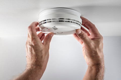 Two male hands holding the edges of a smoke alarm, which is being fitted on the ceiling of a domestic building