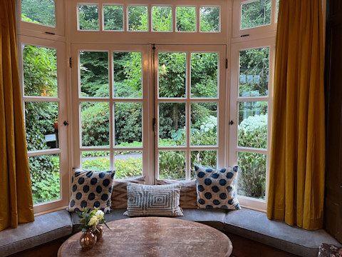 A beige bay window seat forms a cosy window nook with blue and beige cushions. The edge of a brown table can be seen in the foreground. Through the white-framed window is lush greenery outside. Yellow curtains frame the tranquil, inviting setting.