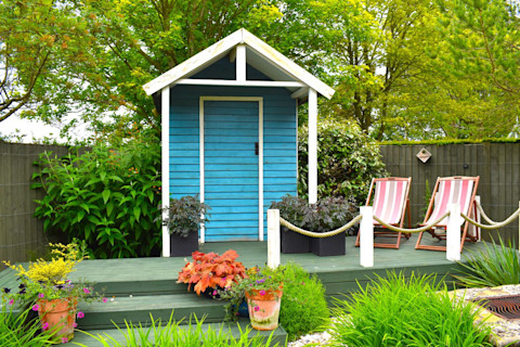 This image shows a small wooden tool shed painted bright blue with a decking area complete with two deck chairs.