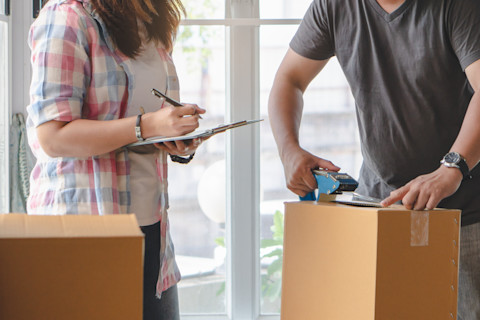 This image shows a couple packing a cardboard box. A woman is checking things off on a clipboard ensuring nothing is forgotten.