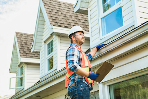 A construction worker in a hard hat and safety vest inspects the roof of a three storey house exterior with a clipboard, conveying professionalism and focus. The background of the image shows three windows on the top floor of the house.