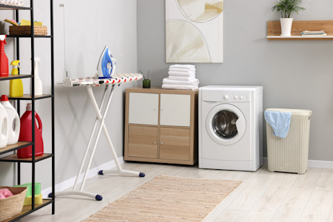 This image shows a utility room complete with a washing machine, laundry basket, a cupboard unit with folded white towels, an ironing board with an iron a shelving unit with cleaning products. The walls are painted a light grey colour and there is a small rattan rug on the floor.