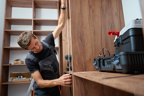 This image shows a carpenter using a measuring tape while fitting mid-tone wood wardrobes in a room. He is wearing overalls and there is a tool kit in the frame.