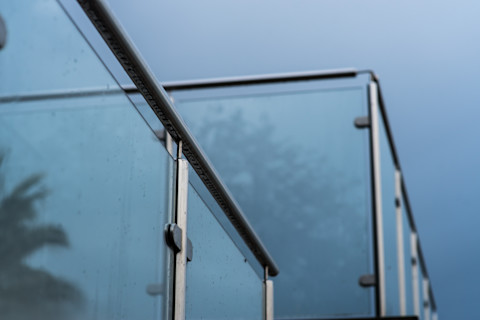 Close-up of a glass balustrade installed along a staircase or balcony. The image shows clear glass panels supported by metal or glass fittings, with a modern and sleek design. The background includes the surrounding area, such as stairs or a balcony.