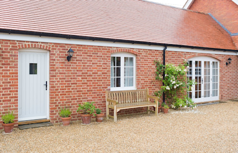 House extension providing a single storey granny annexe. It is a red brick building with a white entry door, patio doors, and window. Beneath the window is a wooden bench on a gravel lawn. Several potted plants and a climbing plant are outside.