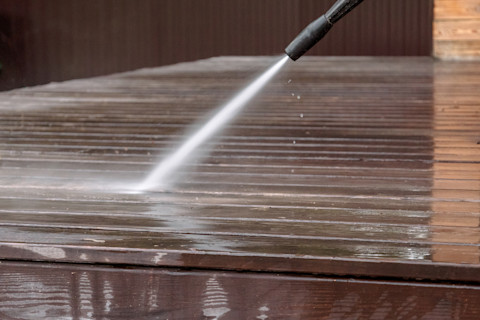 A person uses a pressure washer to clean the surface of a wooden deck, already half cleaned, removing dirt and debris with a strong stream of water.