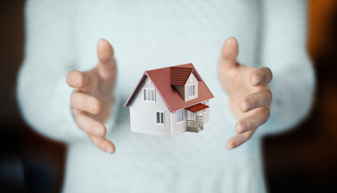 A photo depicting an illusion of a person holding a small, detailed model of a two-story house with a red roof, seemingly suspended in mid-air between their hands. The individual's body is not in focus, emphasizing the house model. They wear a light-colored sweater, and the background is a soft, blurry neutral shade, highlighting the sharp details of the tiny house.